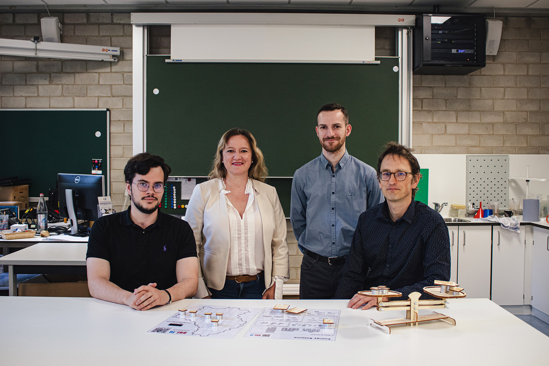 Un groupe de 3 hommes et une femme dans unse salle de classe qui ressemble un laboratoire scientifique. Ils sont debout devant un tableau et regardent droit dans la caméra.