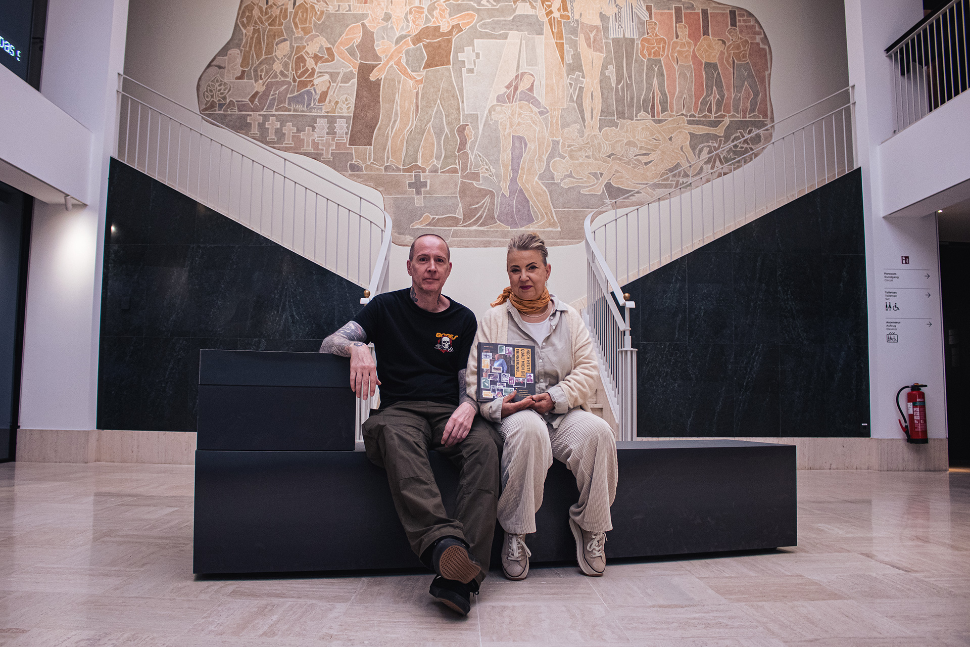 un homme et une femme sont assis sur un banc dans l'entrée du Musée National de la Résistance et des Droits Humains