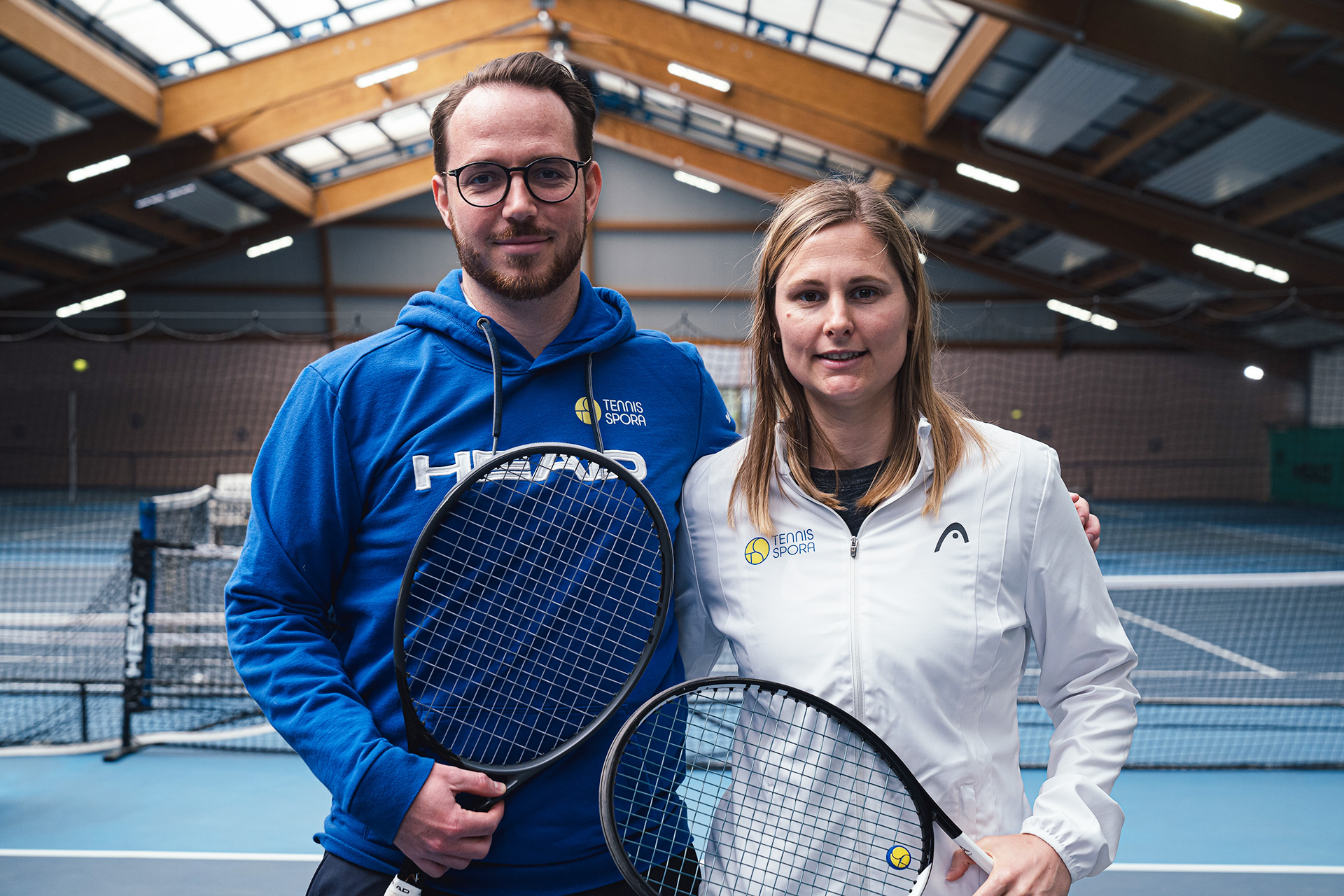 Un homme et une femme posent pour la photo dans un hall de tennis. L’homme, situé à gauche, tient une raquette.